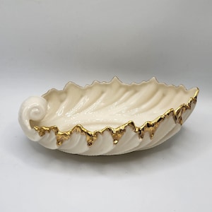 May include: A white ceramic leaf-shaped serving dish with a gold rim. The dish has a textured surface and a curled handle.