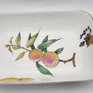 May include: Rectangular white ceramic serving dish with a painted design of peaches, grapes, and leaves. The dish has a gold-coloured trim around the edges. The peaches are yellow and pink, and the grapes are purple.