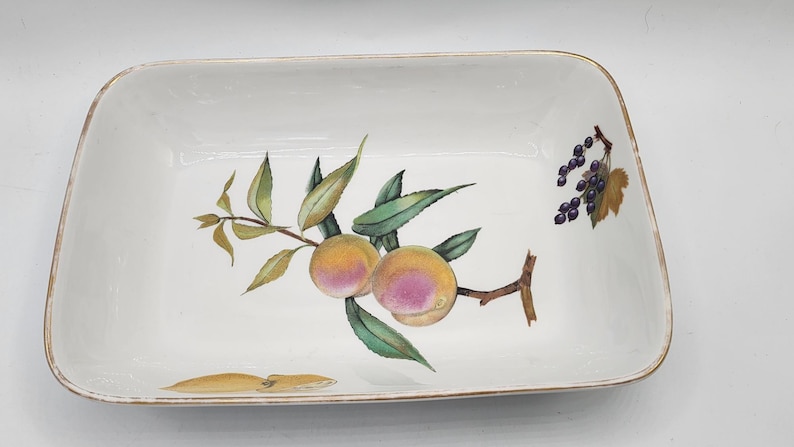 May include: Rectangular white ceramic serving dish with a painted design of peaches, grapes, and leaves. The dish has a gold-coloured trim around the edges. The peaches are yellow and pink, and the grapes are purple.