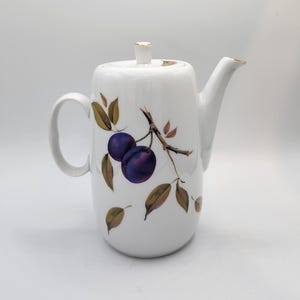 Pode incluir: Bule de cerâmica branco com uma alça e bico curvos. O bule é decorado com um desenho de ramo de ameixa com duas ameixas roxas e folhas marrons e verdes. A tampa e o bico têm detalhes dourados.