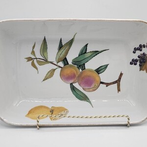 May include: Rectangular white ceramic serving dish with a painted fruit design. The dish features a branch with peaches and leaves, along with lemons and berries. The rim has a gold trim. The dish is displayed on a metal stand.