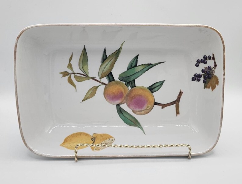 May include: Rectangular white ceramic serving dish with a painted fruit design. The dish features a branch with peaches and leaves, along with lemons and berries. The rim has a gold trim. The dish is displayed on a metal stand.