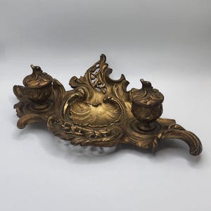 May include: An ornate gold-colored desk inkstand featuring a central shell-shaped pen tray flanked by two lidded inkwells. The piece is crafted with intricate scrolling leaf and vine motifs throughout its surface, finished in a weathered metallic gold.