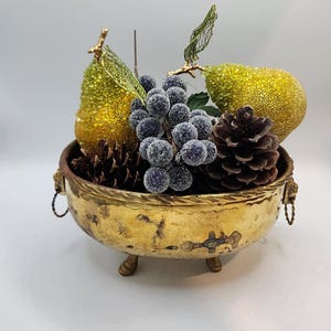 Może przedstawiać: Ozdobna, złota miska wypełniona sztucznymi owocami i szyszkami. Misa ma teksturowaną powierzchnię i jest podparta trzema małymi nóżkami. Kompozycja zawiera dwie błyszczące gruszki, kiść winogron i dwie szyszki.
