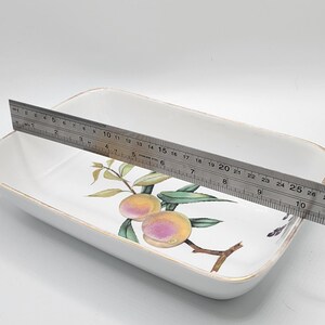 May include: Rectangular white ceramic serving dish with a peach and leaf design. The dish is approximately 25 cm long. A stainless steel ruler is placed across the dish.