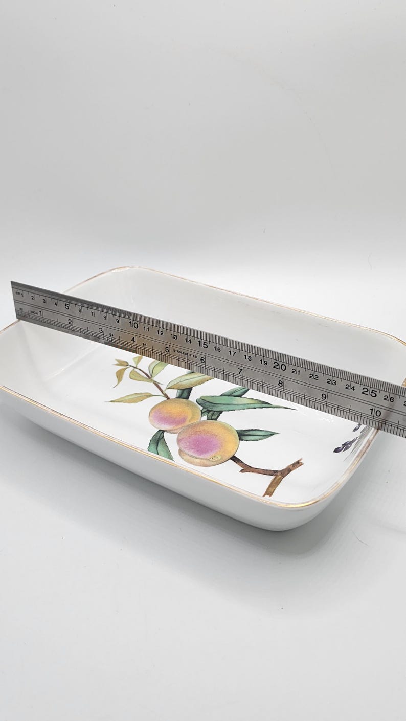 May include: Rectangular white ceramic serving dish with a peach and leaf design. The dish is approximately 25 cm long. A stainless steel ruler is placed across the dish.