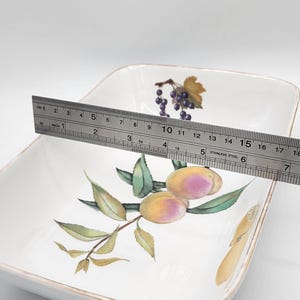 May include: White square serving dish with a gold rim, decorated with a fruit design. The dish is approximately 18 cm wide. The fruit design includes grapes and plums.
