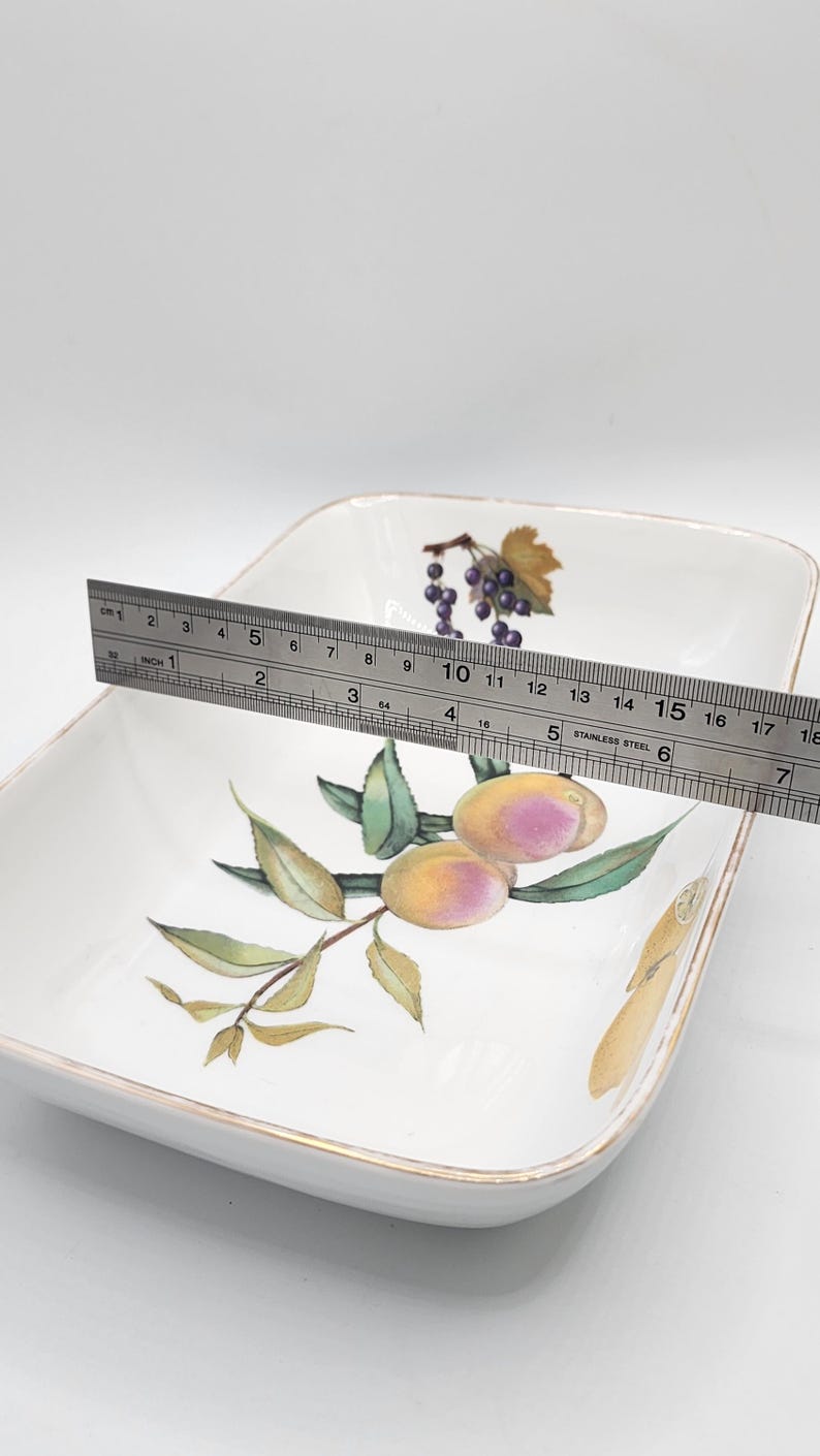 May include: White square serving dish with a gold rim, decorated with a fruit design. The dish is approximately 18 cm wide. The fruit design includes grapes and plums.