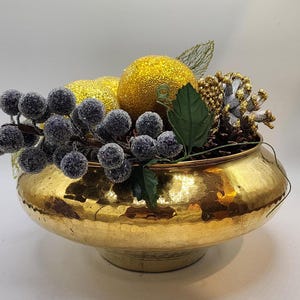 Puede incluir: Un cuenco de metal dorado martillado lleno de elementos decorativos. El cuenco contiene dos esferas doradas cubiertas de purpurina, racimos de bayas azul oscuro, hojas verdes y acentos de piñas. El cuenco se asienta sobre una pequeña base redonda.