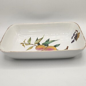 May include: Rectangular white ceramic serving dish with rounded corners. The dish features a painted design of peaches and grapes with green leaves. The rim has a gold trim. A decorative piece for serving food.