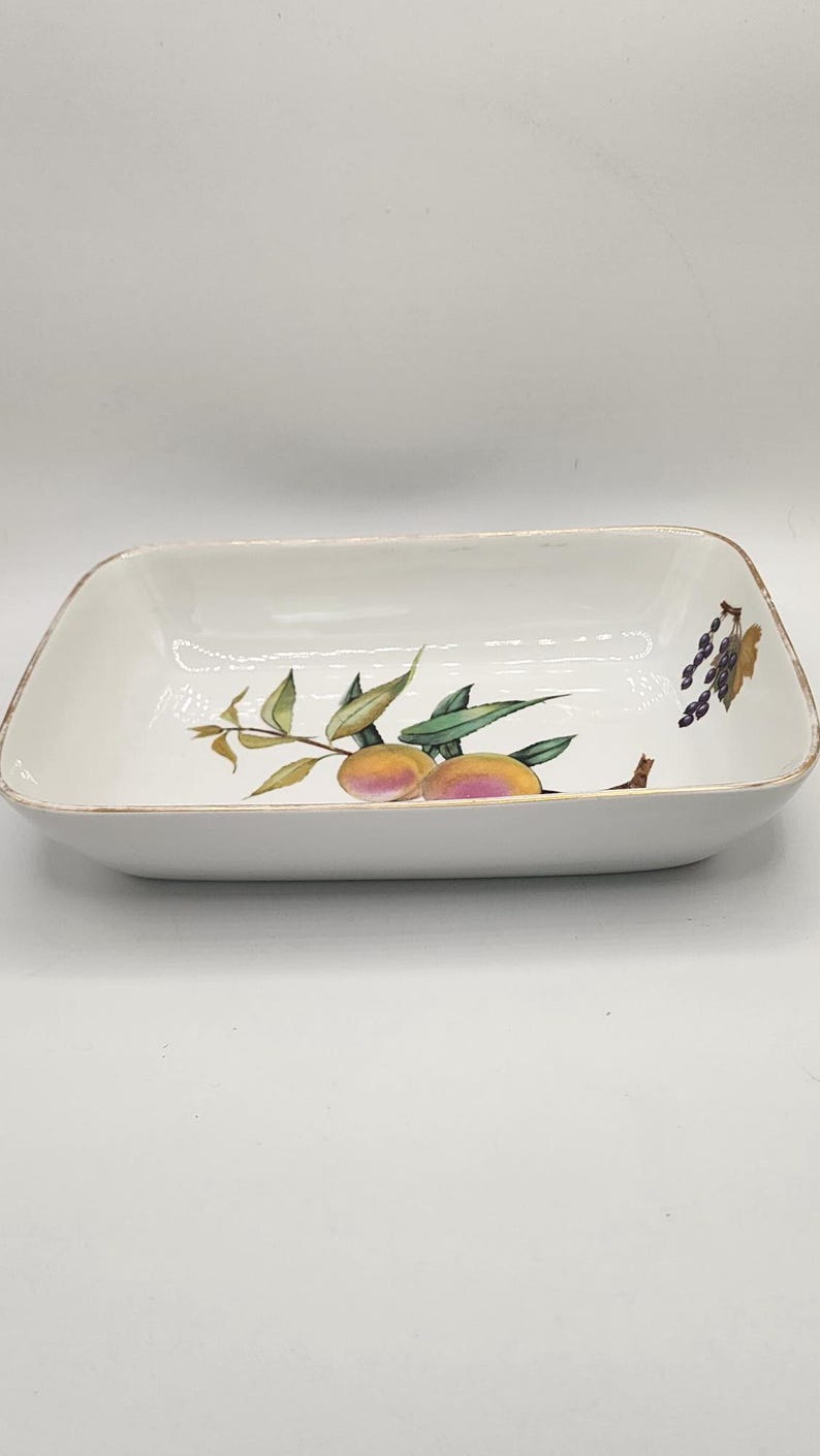 May include: Rectangular white ceramic serving dish with rounded corners. The dish features a painted design of peaches and grapes with green leaves. The rim has a gold trim. A decorative piece for serving food.