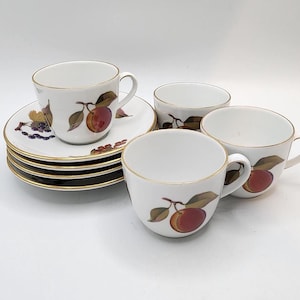 Pode incluir: Um conjunto de chávenas e pires de porcelana branca. Cada chávena e pires apresenta uma borda dourada e um desenho de uma ameixa vermelha com folhas verdes. O conjunto inclui quatro chávenas e uma pilha de pires.
