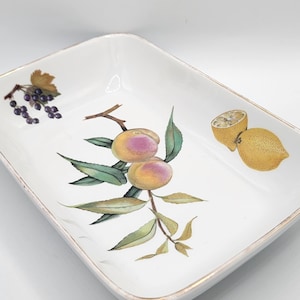 Puede incluir: Plato rectangular de cerámica blanca con borde dorado. El plato está decorado con ilustraciones de melocotones, uvas y limones. Las ilustraciones de frutas son en tonos amarillos, rosas y verdes.