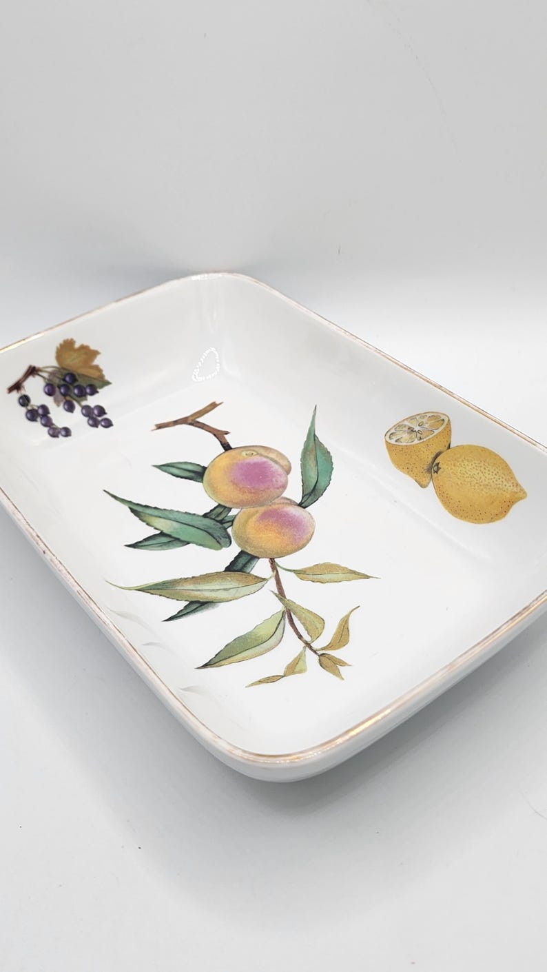 May include: Rectangular white ceramic serving dish with a gold rim. The dish is decorated with illustrations of peaches, grapes, and lemons. The fruit illustrations are in shades of yellow, pink, and green.
