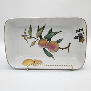 May include: White rectangular ceramic serving dish with a hand-painted fruit design. The dish features a branch with peaches and green leaves, and a smaller branch with dark berries. The dish is supported by a gold-coloured metal stand.