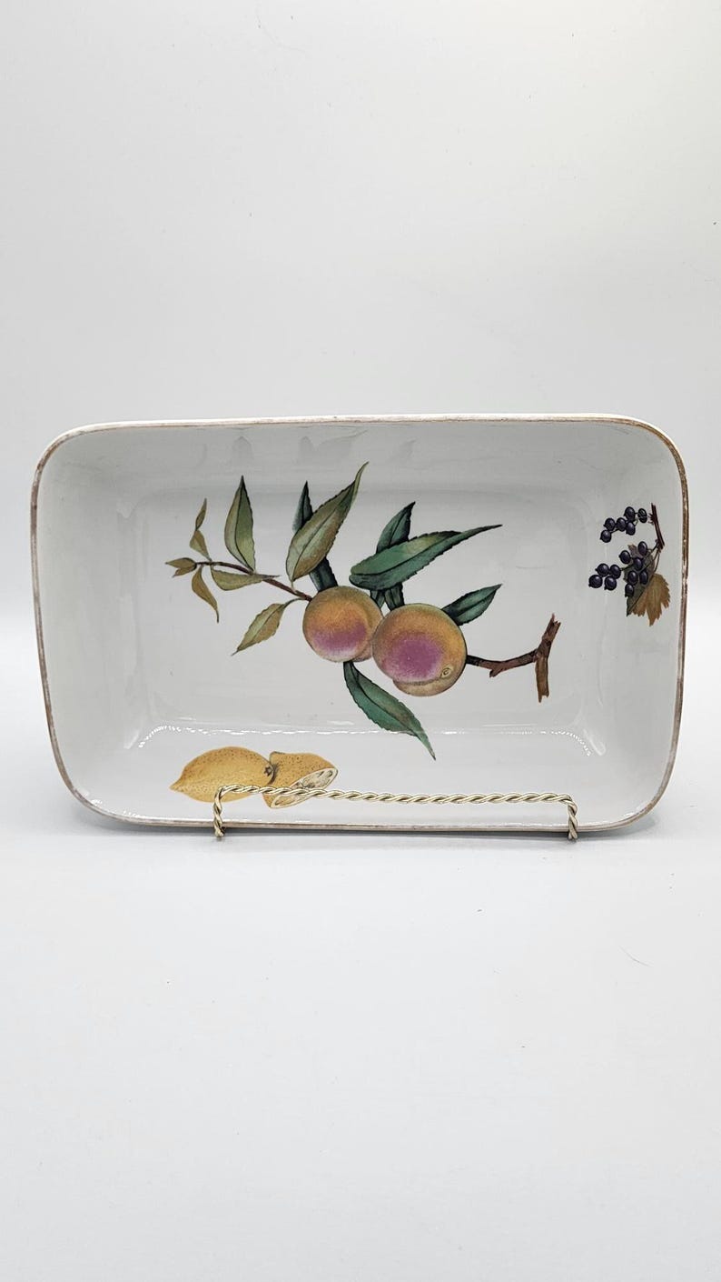 May include: White rectangular ceramic serving dish with a hand-painted fruit design. The dish features a branch with peaches and green leaves, and a smaller branch with dark berries. The dish is supported by a gold-coloured metal stand.