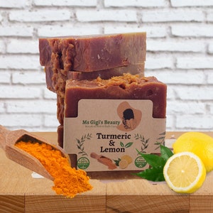 Peut inclure: Une pile de savons bruns et orange avec une étiquette qui indique "Ms Gigi's Beauty Botanical Hair & Body Care Curcuma & Citron 100% Naturel Fait à la Main". Les savons sont posés sur une surface en bois avec une cuillère de poudre de curcuma et deux citrons.
