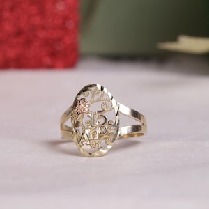 May include: Gold-toned ring with an oval filigree design. The ring features a floral accent and the inscription "15 Años". The band splits into two parallel lines on either side of the oval.