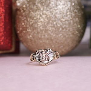 Puede incluir: Un anillo de oro en forma de coraz&oacute;n con un coraz&oacute;n blanco en el centro que presenta el n&uacute;mero "15" en su interior. El anillo est&aacute; sobre una superficie rosa con adornos brillantes rojos, dorados y verdes en el fondo.