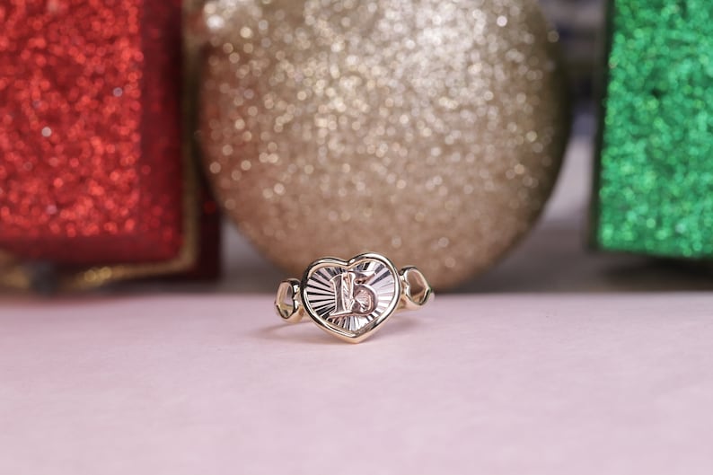 Puede incluir: Un anillo de oro en forma de coraz&oacute;n con un coraz&oacute;n blanco en el centro que presenta el n&uacute;mero "15" en su interior. El anillo est&aacute; sobre una superficie rosa con adornos brillantes rojos, dorados y verdes en el fondo.