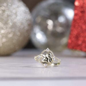 K&ouml;nnte beinhalten: Ein goldener Ring mit einem kleinen Engelsmotiv und klaren Steinen. Der Ring liegt auf einer wei&szlig;en Oberfl&auml;che.