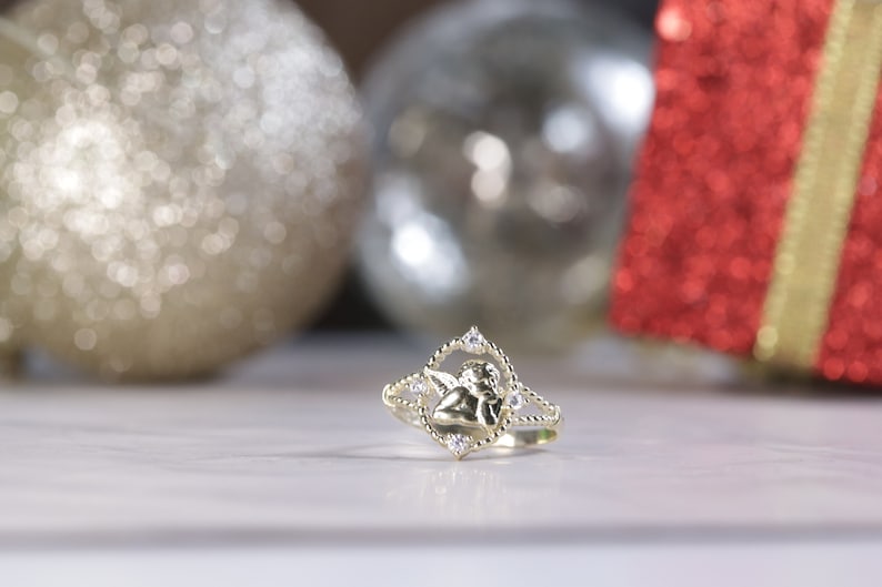 K&ouml;nnte beinhalten: Ein goldener Ring mit einem kleinen Engelsmotiv und klaren Steinen. Der Ring liegt auf einer wei&szlig;en Oberfl&auml;che.