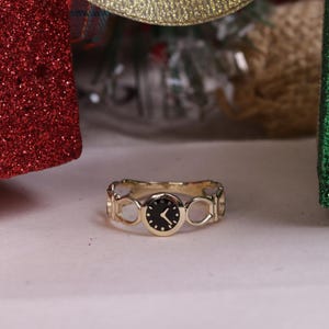 May include: A gold-colored ring with a black watch face. The ring has a unique design with open circular cutouts. The watch face has white hands and markers. The ring is displayed in front of red and green glittery gift boxes.