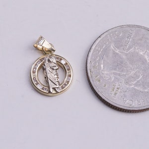 May include: A gold pendant with a small, white, detailed image of a saint inside a circle of small, clear stones. The pendant is about the size of a US quarter.