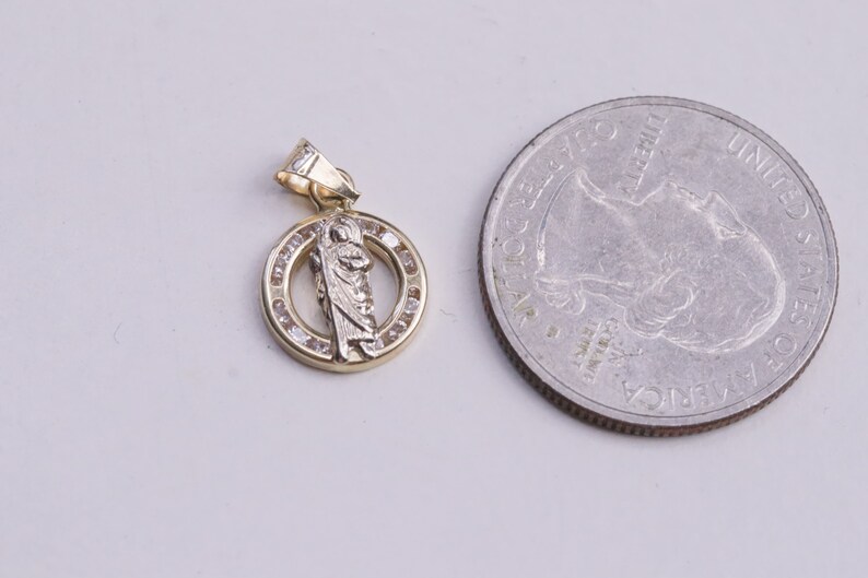 May include: A gold pendant with a small, white, detailed image of a saint inside a circle of small, clear stones. The pendant is about the size of a US quarter.