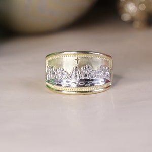 May include: A gold and silver ring with a detailed engraving of a religious scene. The ring is wide and has a rounded shape.