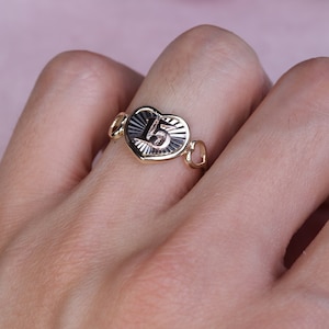 Puede incluir: Un anillo de oro en forma de coraz&oacute;n con el n&uacute;mero "15" grabado en &eacute;l. El anillo tiene un delicado dise&ntilde;o de cadena.