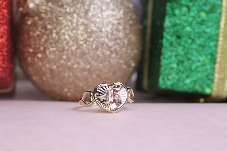 Puede incluir: Un anillo de oro en forma de coraz&oacute;n con el n&uacute;mero "15" grabado en el interior. El anillo est&aacute; rodeado por dos corazones m&aacute;s peque&ntilde;os a cada lado.