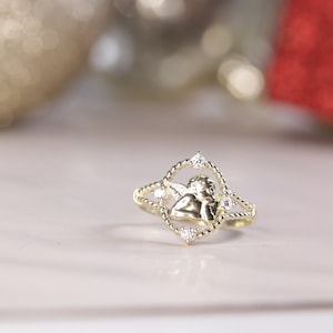 K&ouml;nnte beinhalten: Ein goldener Ring mit einem kleinen Engelsmotiv und klaren Zirkonia-Steinen.