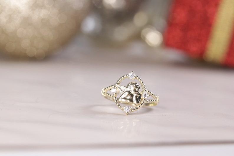K&ouml;nnte beinhalten: Ein goldener Ring mit einem kleinen Engelsmotiv und klaren Zirkonia-Steinen.