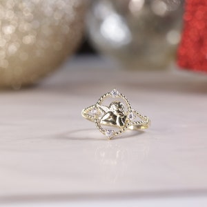 K&ouml;nnte beinhalten: Ein goldener Ring mit einem kleinen Engelsmotiv und klaren Steinen. Der Ring liegt auf einer wei&szlig;en Oberfl&auml;che.