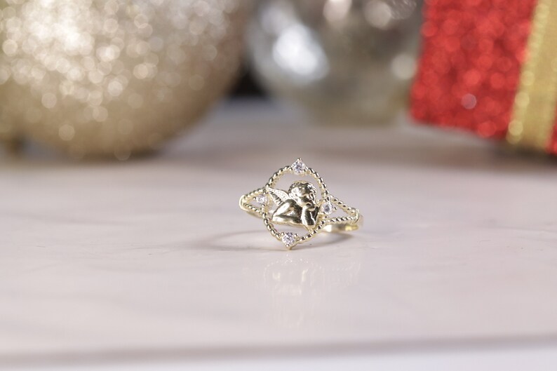 K&ouml;nnte beinhalten: Ein goldener Ring mit einem kleinen Engelsmotiv und klaren Steinen. Der Ring liegt auf einer wei&szlig;en Oberfl&auml;che.