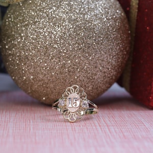 May include: A gold ring with the number 15 in the center, flanked by two small clear stones. The ring has an ornate, openwork design. The background includes a gold glitter ornament, a green gift box, and a red gift box.
