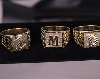May include: Three gold signet rings with the letters L, M, and D engraved on the top. The rings have a textured design.