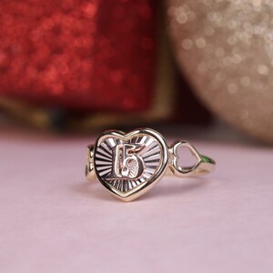 Puede incluir: Un anillo de oro en forma de coraz&oacute;n con el n&uacute;mero "15" grabado en el interior. El anillo tiene un delicado dise&ntilde;o de coraz&oacute;n abierto en el lateral.