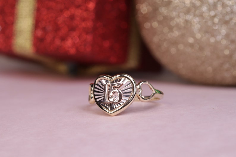 Puede incluir: Un anillo de oro en forma de coraz&oacute;n con el n&uacute;mero "15" grabado en el interior. El anillo tiene un delicado dise&ntilde;o de coraz&oacute;n abierto en el lateral.