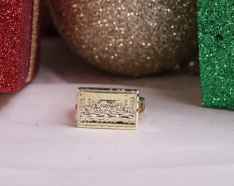 May include: A gold-colored ring with a rectangular design featuring a raised image of a scene with many people. The ring is on a white surface.