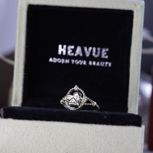 K&ouml;nnte beinhalten: Ein silberner Ring mit einem herzf&ouml;rmigen Design und kleinen, klaren Steinen. Der Ring befindet sich in einer wei&szlig;en Schachtel mit schwarzem Samtbezug. Die Schachtel tr&auml;gt den Text "HEAVUE ADORN YOUR BEAUTY" auf dem Deckel.