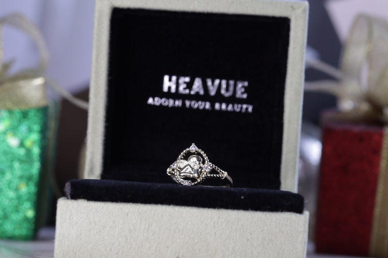 K&ouml;nnte beinhalten: Ein silberner Ring mit einem herzf&ouml;rmigen Design und kleinen, klaren Steinen. Der Ring befindet sich in einer wei&szlig;en Schachtel mit schwarzem Samtbezug. Die Schachtel tr&auml;gt den Text "HEAVUE ADORN YOUR BEAUTY" auf dem Deckel.