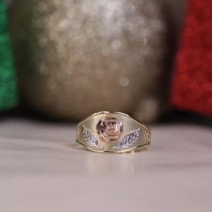 May include: A gold ring with a rose design in the center. The rose is made of three different shades of gold, with the center being a darker shade of gold.