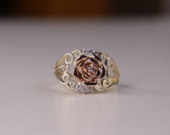 May include: A gold-tone ring featuring a rose design with a two-tone rose gold and gold flower center. The ring has a decorative filigree design with small, clear accent stones. The ring is a piece of jewelry.