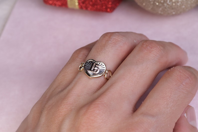 Puede incluir: Anillo de oro en forma de coraz&oacute;n con el n&uacute;mero "15" grabado en el interior. El anillo est&aacute; en un dedo.