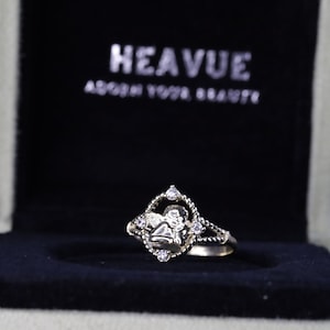 K&ouml;nnte beinhalten: Ein goldener Ring mit einer diamantf&ouml;rmigen Fassung, die ein Engelsmotiv zeigt. Der Ring befindet sich in einer schwarzen Samtbox mit den Worten "Heavue Adorn Your Beauty" auf dem Deckel.
