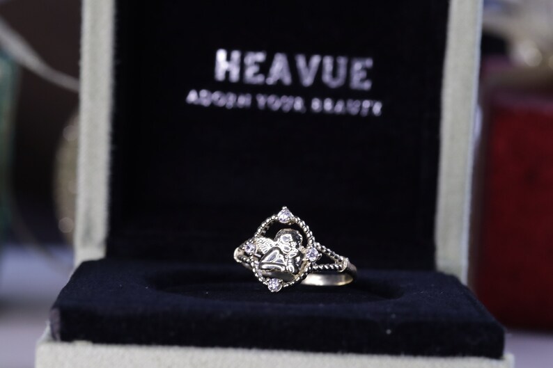 K&ouml;nnte beinhalten: Ein goldener Ring mit einer diamantf&ouml;rmigen Fassung, die ein Engelsmotiv zeigt. Der Ring befindet sich in einer schwarzen Samtbox mit den Worten "Heavue Adorn Your Beauty" auf dem Deckel.