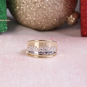 Può includere: Un anello in oro e argento con un designo in rilievo di una croce e un gruppo di persone. L'anello è su una superficie rosa.