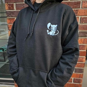 Könnte beinhalten: Ein schwarzer Kapuzenpullover mit einem hellblauen gestickten Comic-Charakter auf der Vorderseite. Der Charakter hat große Augen und einen eingerollten Schwanz.